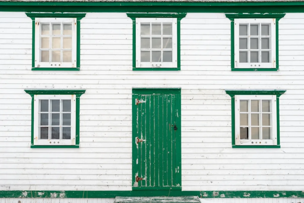 The exterior wall of a vintage white wooden building with bright green trim. There's a small wooden door and three glass windows in the country house. The wall has a textured horizontal clapboard.