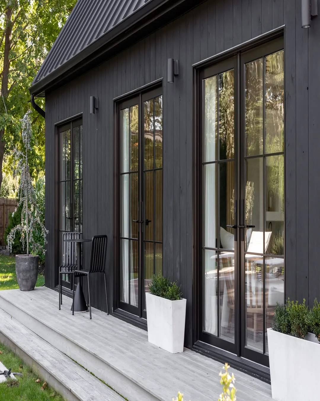 Black-framed glass doors along dark vertical siding, with planters on a wood deck.