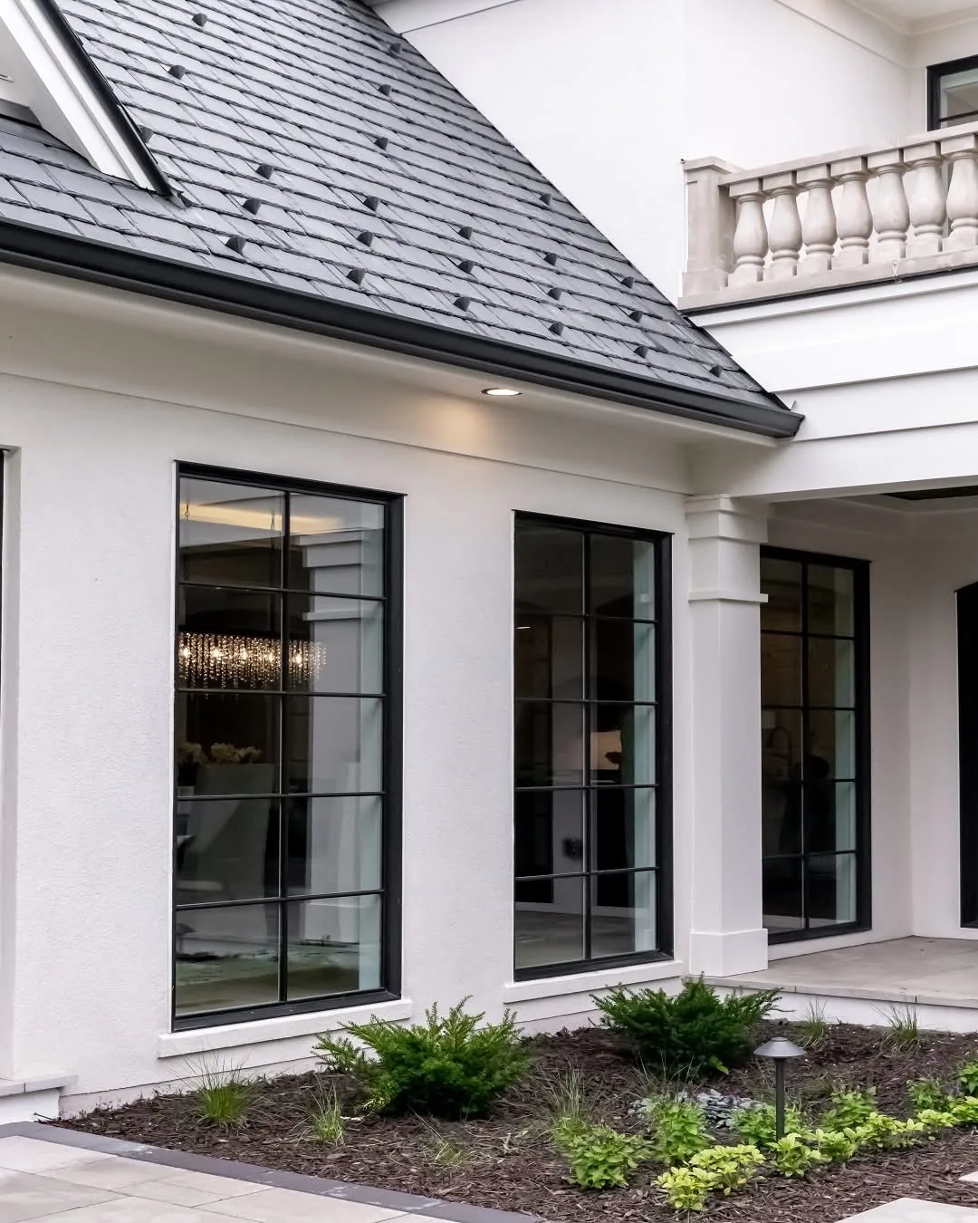 Dark sloped roof above black-framed windows on a white home with a landscaped walkway.