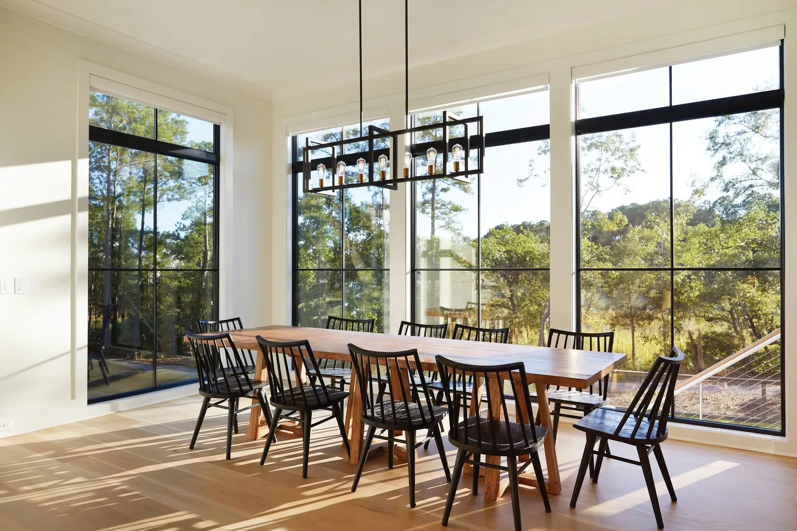 Dining room with a long wooden table and tall black-framed windows overlooking trees.