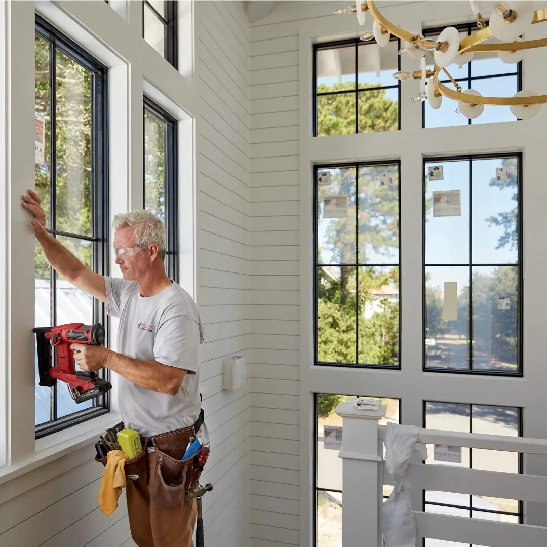Person working on interior trim beside tall black-framed windows with a handheld power tool.