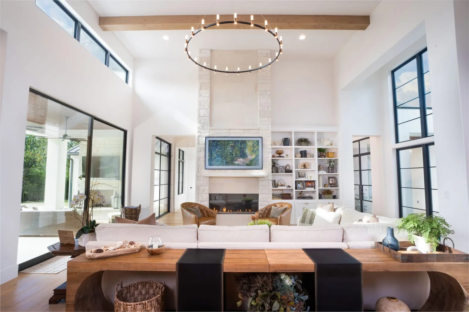 Living room with tall black-framed windows, a stone fireplace, white seating, and a round chandelier.